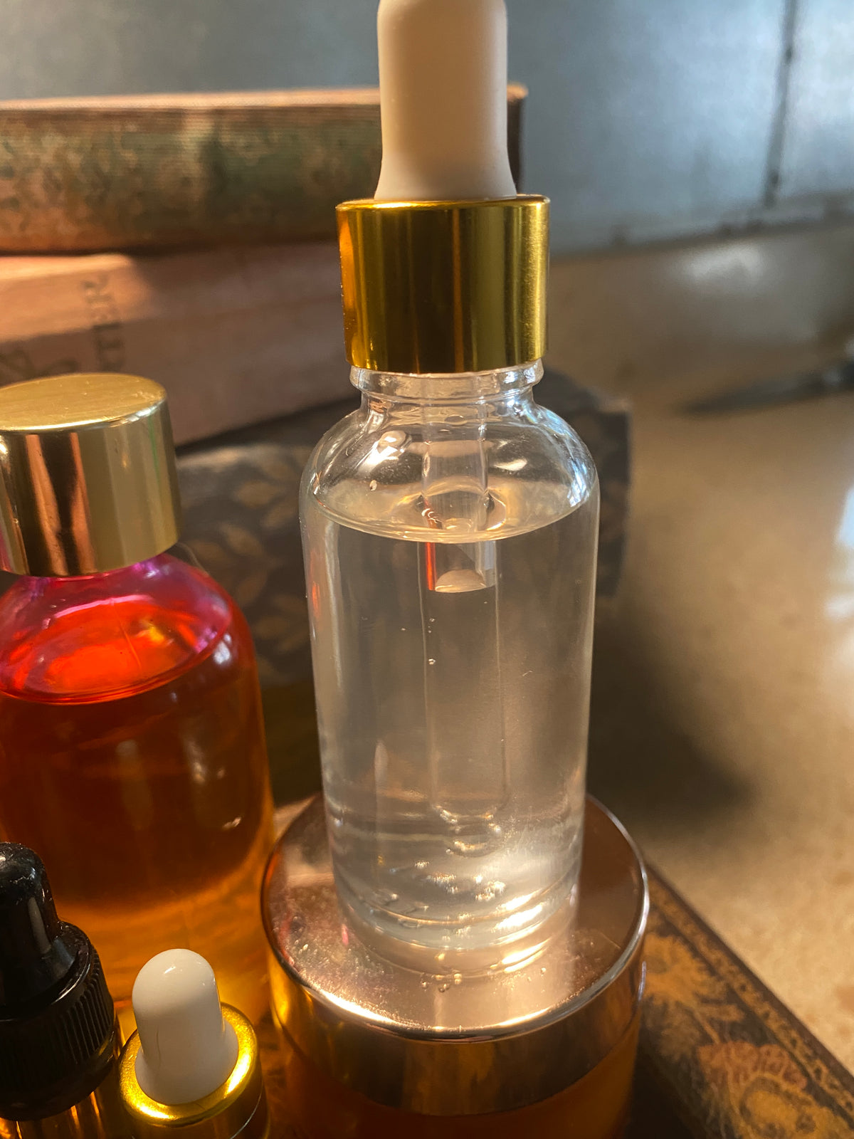 A clear glass dropper bottle filled with clear liquid, featuring a shiny gold collar and a white rubber bulb, resting on a metallic gold base next to other cosmetic bottles.
